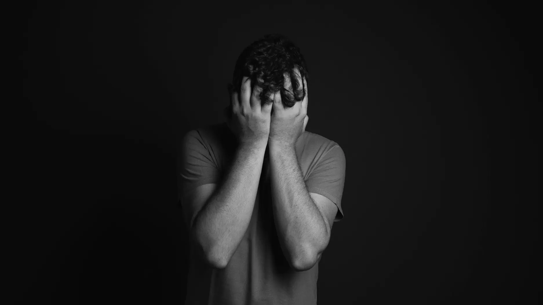 Persoană acoperindu-și fața cu mâinile, exprimând suferință sau anxietate, pe fundal întunecat.